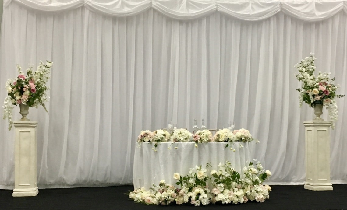 This wedding staging is created by Fabulous Functions UK and  included a sweetheart table decorated with an abundance of flowers. There were flowers on the table, flowers surrounding the table and two floral arrangements on pedestals either side if the sweetheart table