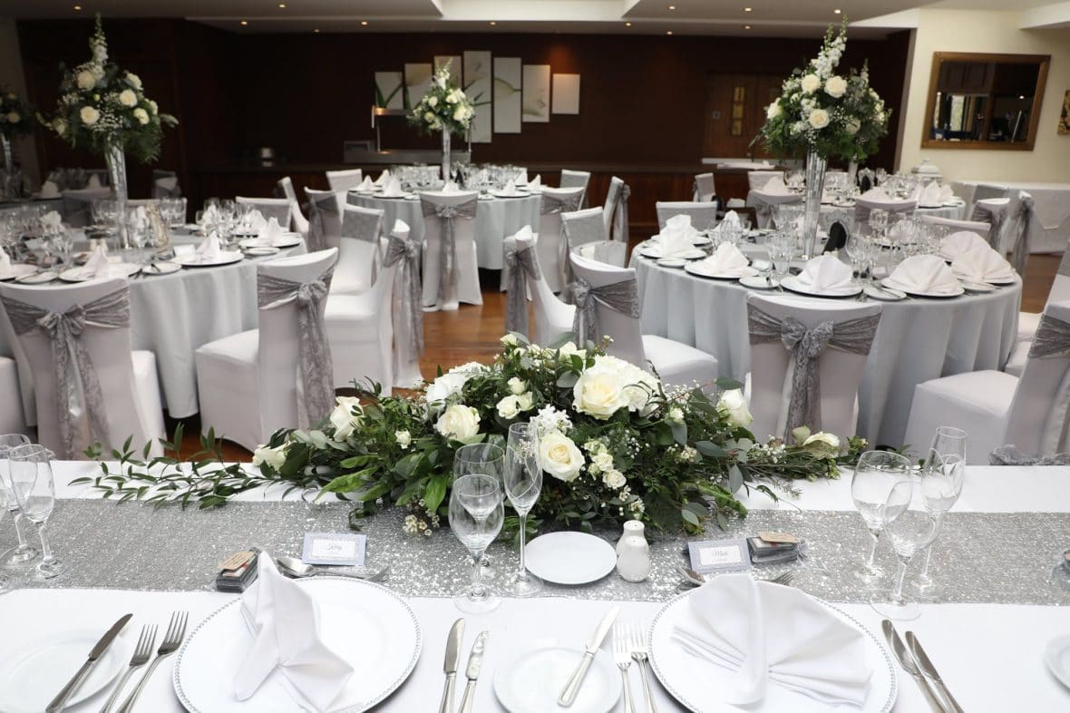 A Silver & Grey Theme wedding decor featuring silver grey table cloth, silver satin and silver lace sashes white charger plates with diamante rims. Blush pinlk flower wall and white florals with greenery-Fabulous Function UK