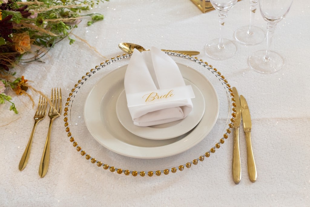 Place setting of gold beaded edged glass charger plate with acrylic guest name plates - guest names written in gold coloured font and displayed on a  white folded  napkin.