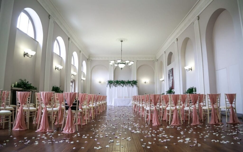 Rose gold chiffon vertical drops serve to decorate the chiavari chairs in the Assembly Rooms of the Kings Head Hotel in Cirencester. The backdrop was created with a white voile draped panel and green foliage for the top border decoration. Styling by Fabulous Functions UK