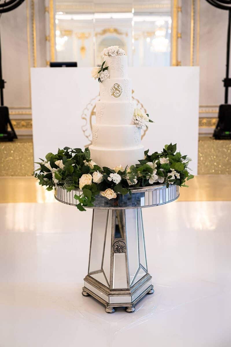 Wedding cake displayed on a mirrored cake table with green and white garland decor